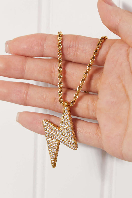 Hand holding Brass Lightning Pendant Necklace with twisted chain and sparkling cubic zirconia detail in flat lay style.