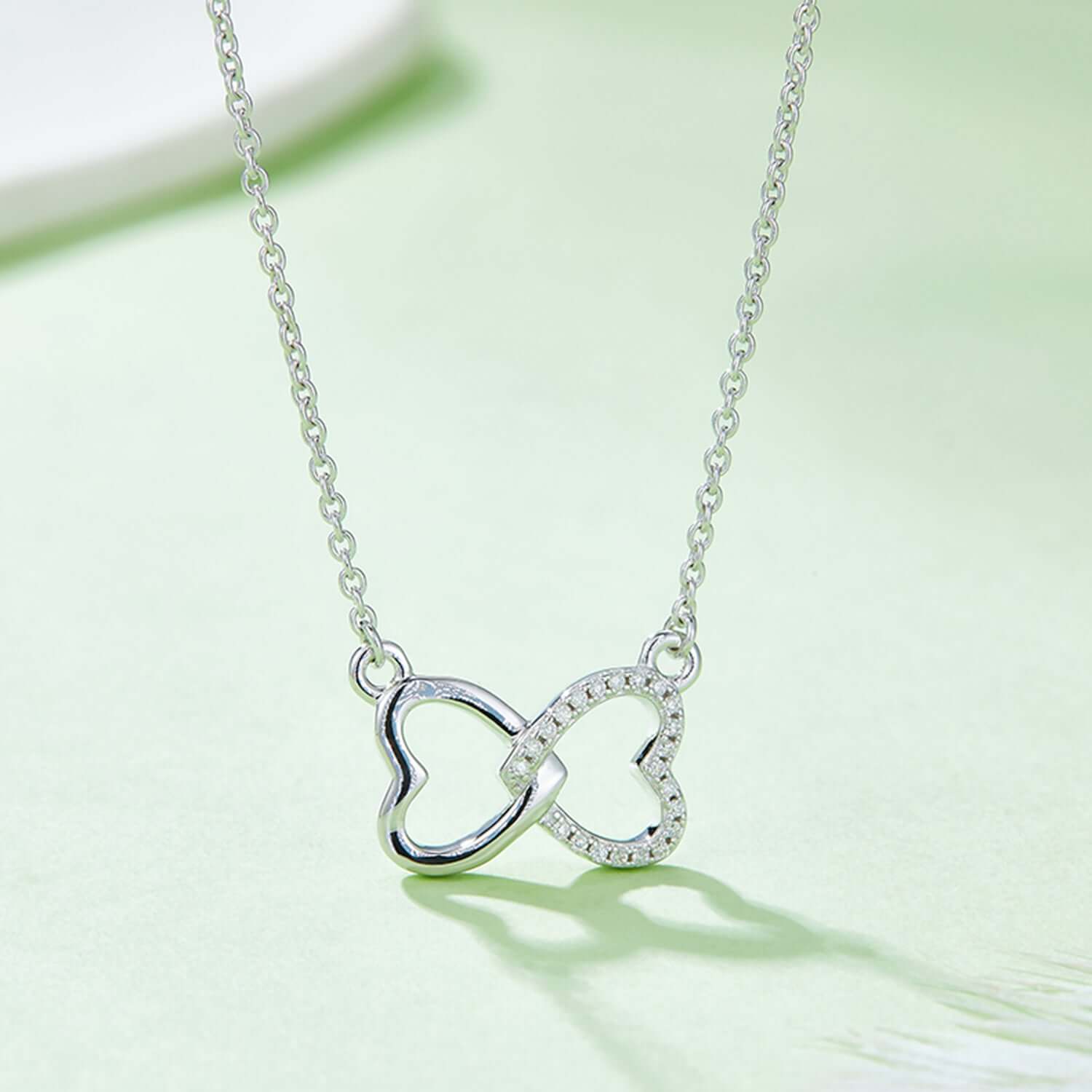 Moissanite 925 sterling silver heart bow necklace on light green background, featuring interlocking heart design with a delicate chain.