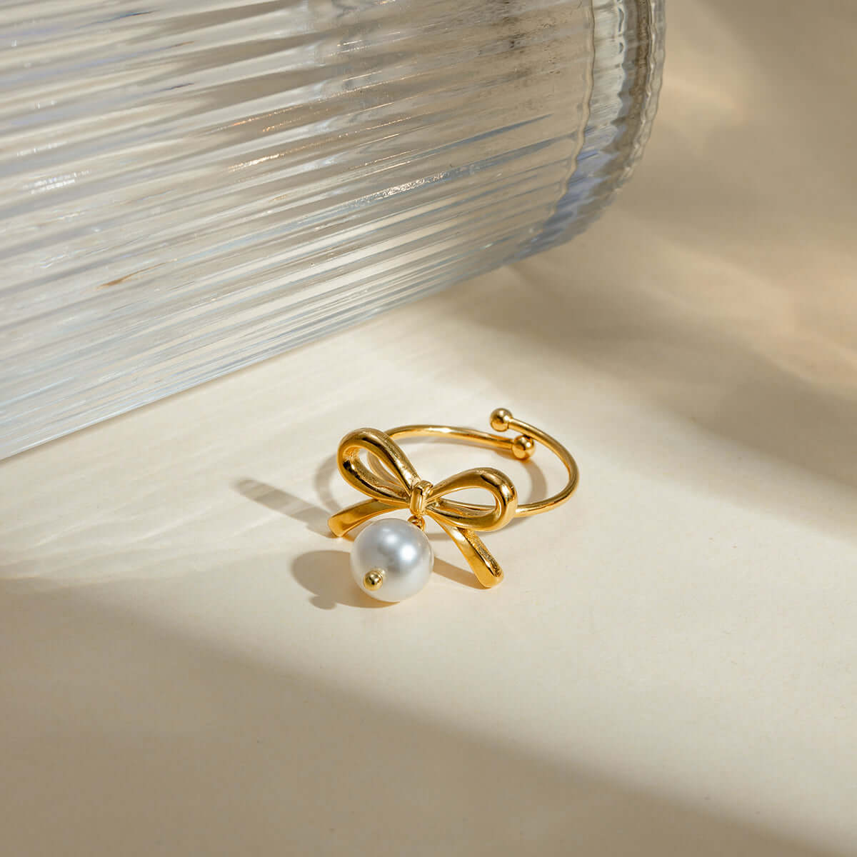 Stainless steel pearl bow ring with synthetic pearl and 18K gold-plated bow design, displayed beside a glass vase.