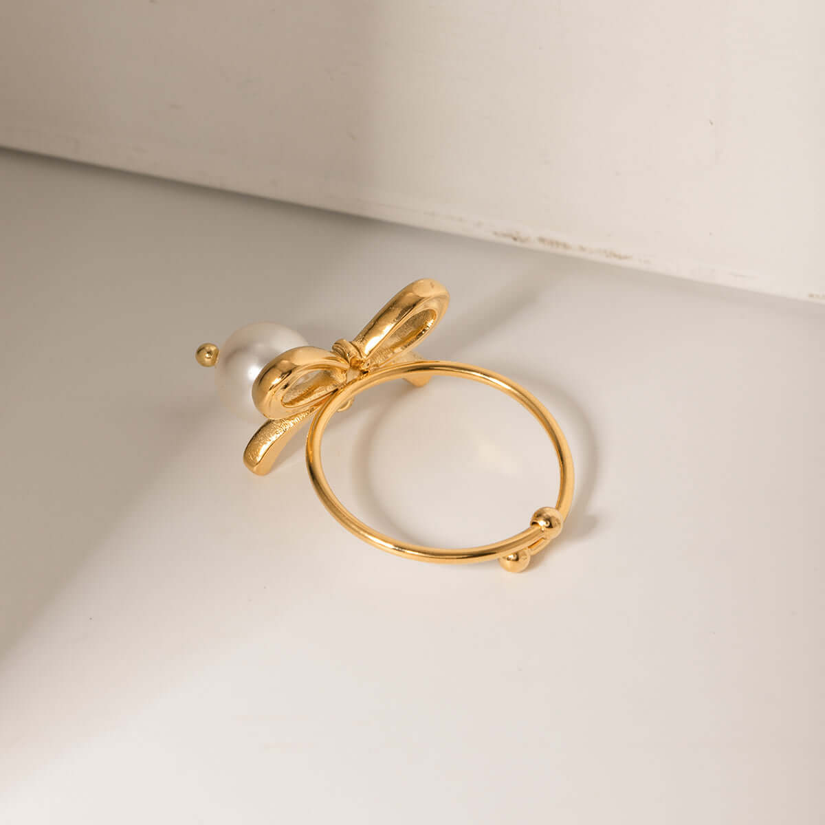 Stainless steel pearl bow ring with 18K gold plating, featuring an elegant synthetic pearl on an adjustable band, displayed on a white surface.