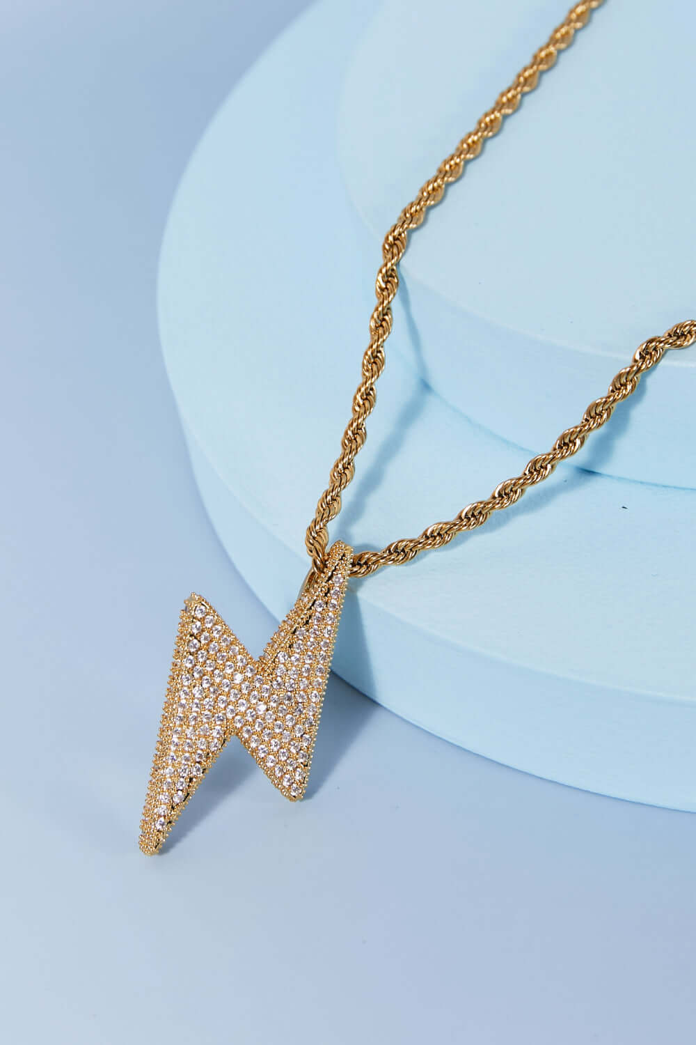 Brass lightning pendant necklace with twisted chain and cubic zirconia detail in flat lay style on a blue background.