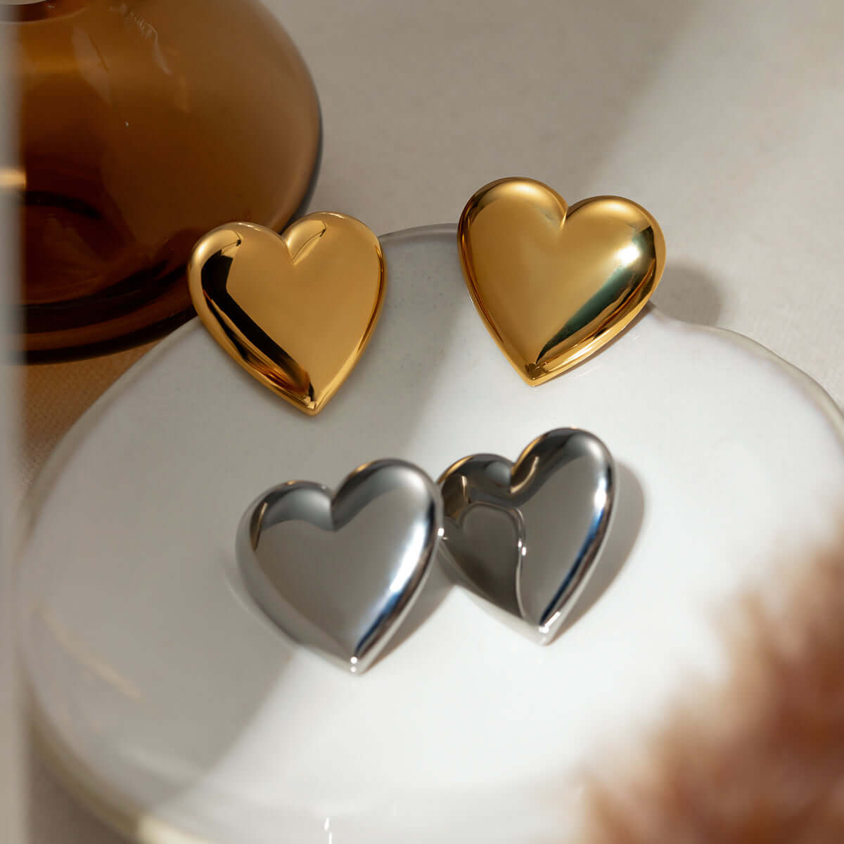 Gold and silver stainless steel heart stud earrings displayed on a white dish, showcasing their reflective surfaces and elegant design.