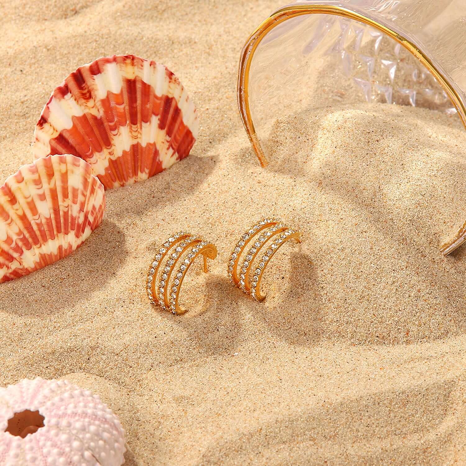 Stainless steel inlaid zircon C-hoop earrings with gold plating on sandy beach with seashells and a transparent glass.