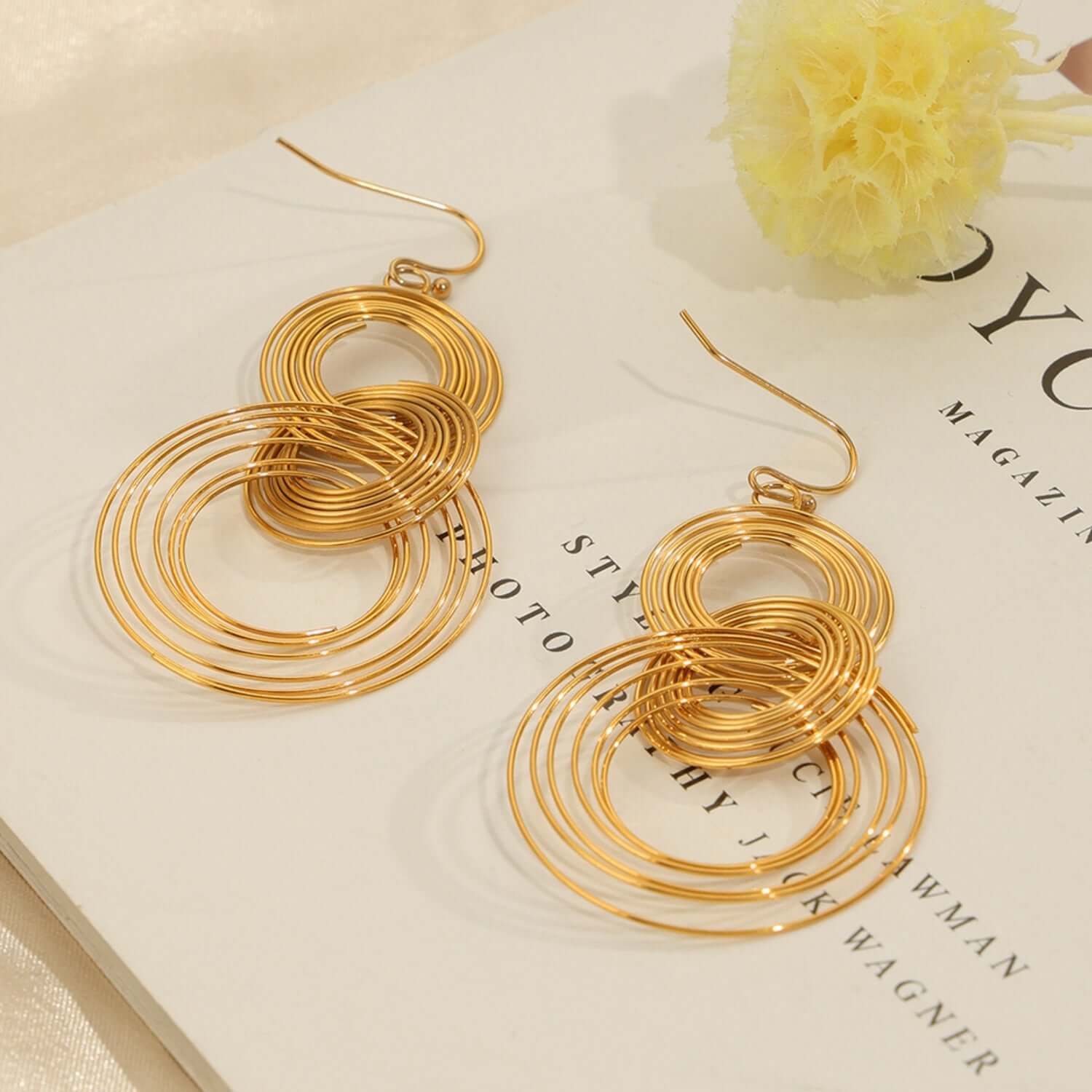 Titanium Steel Multi-Layered Round Shape Earrings in gold-plated finish laid on a book with a yellow flower.