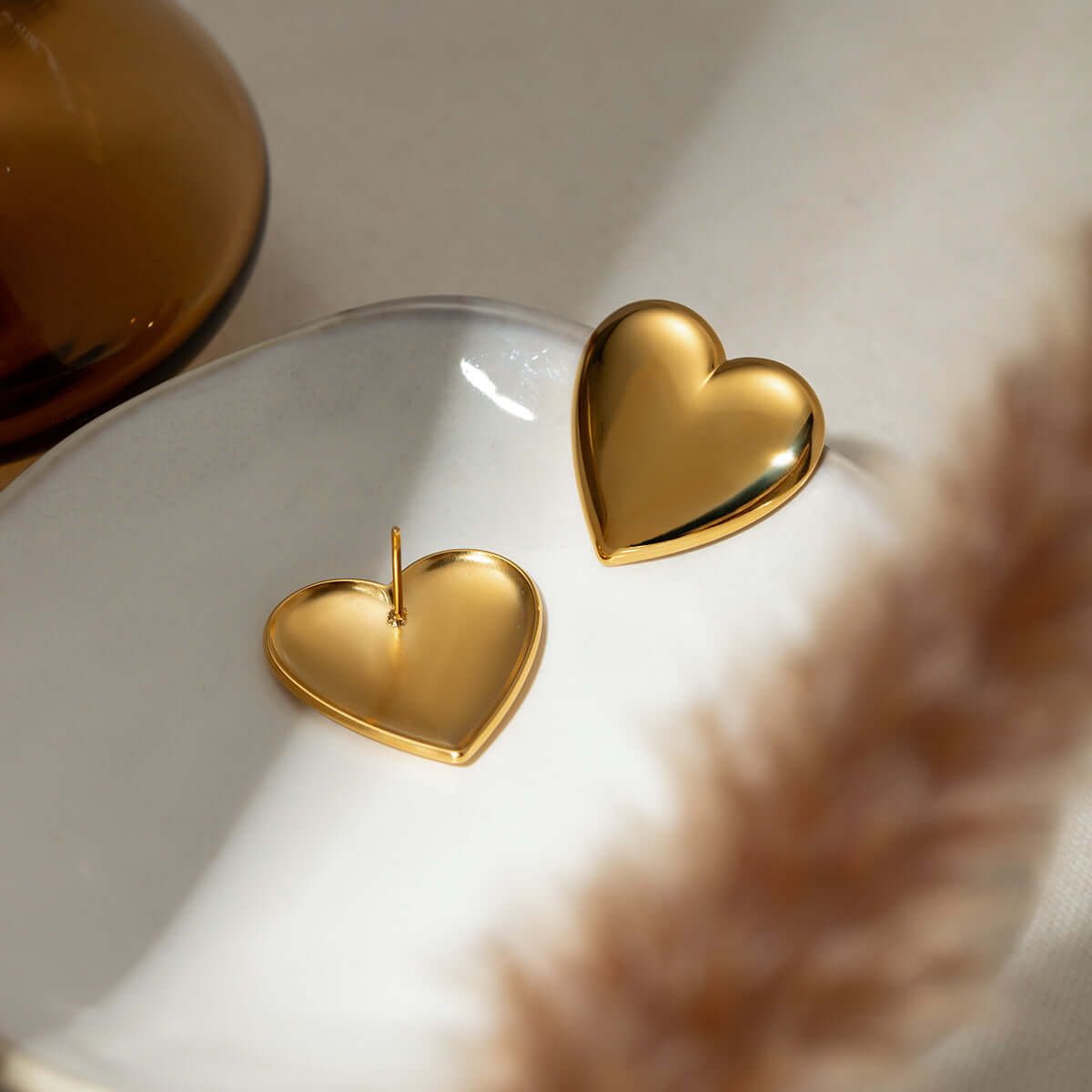 Gold-plated stainless steel heart stud earrings on ceramic dish, showcasing elegant design and reflective surface.