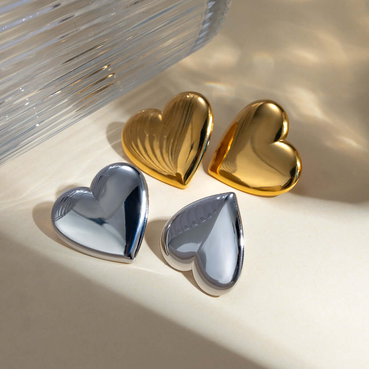 Gold and silver stainless steel heart-shaped stud earrings in sunlight on a beige background.